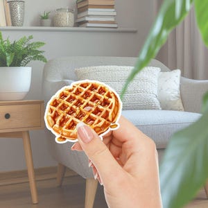 May include: A sticker depicting a golden-brown waffle with syrup. The waffle has a grid pattern and is held in front of a blurred background of a living room with a sofa, plants, and a shelf.