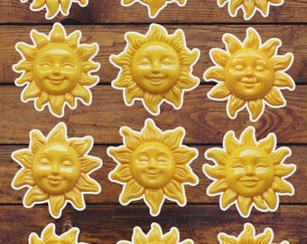 May include: Eleven smiling sun-shaped decorative elements in a collection. Each sun is a bright yellow with a white outline, featuring a smiling face and radiating sun rays. They are arranged on a wooden background.