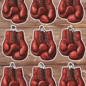 May include: Nine red boxing gloves stickers with black laces and hanging strings. The gloves are in pairs, with a vintage, distressed look. The stickers are arranged on a wooden background.