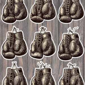 May include: A collection of nine stickers featuring vintage-style boxing gloves. The gloves are depicted in a detailed, sepia-toned illustration, with laces and hanging strings. The stickers are set against a wooden background.