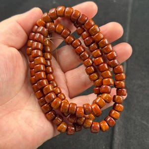 Puede incluir: Un collar de cuentas de madera de color marrón rojizo, cada una con forma rectangular y acabado pulido. Las cuentas están ensartadas, formando un collar o pulsera. Las cuentas se sostienen en una mano.