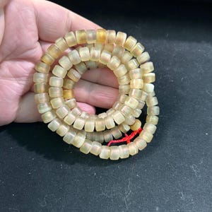 Könnte beinhalten: Eine aufgerollte Halskette aus zahlreichen kleinen, durchscheinenden, rechteckigen Perlen. Die Perlen haben einen hellgelben Farbton mit subtilen Farbvariationen. Eine rote Schnur sichert die Halskette.