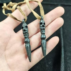 May include: Two dark gray metal pendants, each featuring a skull design at the top and a pointed bottom. They hang from tan leather cords. The pendants are held in a hand.