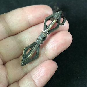 May include: A small, antique bronze vajra, a ritual object with diamond-shaped ends and a central grip. The vajra is held by a hand, showcasing its intricate details and aged patina. The object is likely used for spiritual or ceremonial purposes.
