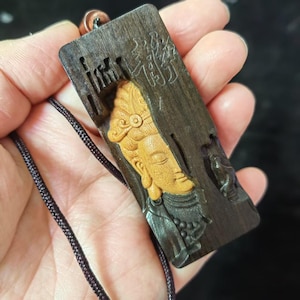 May include: A rectangular wooden pendant with a carved relief of a serene figure. The figure is light brown, set against a dark brown background. The pendant hangs from a black cord.