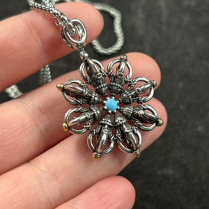 Può includere: Collana con pendente argentato con un design floreale dettagliato. Il pendente ha una pietra centrale turchese e dettagli dorati. Anche la catena della collana è argentata.
