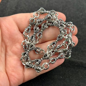 May include: A silver necklace with detailed, ornate patterns. The chain is made up of repeating decorative elements, linked by small rings. The design features a series of stylized, symmetrical shapes, creating a complex and textured look. The necklace is shown against a dark background.
