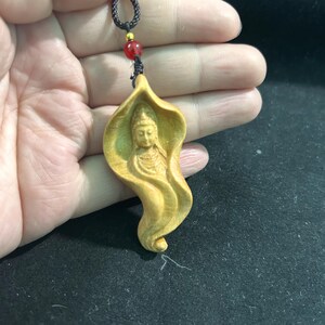 May include: A carved wooden pendant featuring a serene figure within a flowing, leaf-like design. The pendant is a light, natural wood color, with intricate details. It hangs from a black cord with a red bead accent.