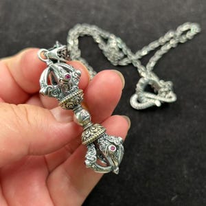 Könnte beinhalten: Silberfarbene Anhänger Halskette mit einem detaillierten, verzierten Vajra-Design. Der Anhänger hat rote Edelsteinakzente und goldfarbene Details. Die Kette ist ebenfalls silberfarben und hat ein strukturiertes Aussehen. Die Halskette wird in einer Hand vor dunklem Hintergrund gehalten.