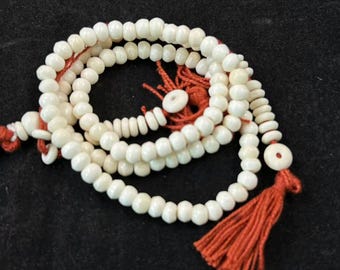 May include: A white beaded necklace with a red tassel and accents. The beads are round and appear to be made of bone or a similar material. The necklace is arranged in a circular pattern against a dark background.