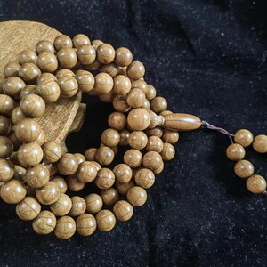 May include: A strand of light brown wooden prayer beads, featuring numerous round beads with visible wood grain. The beads are strung together, with a tassel at the end. The beads are resting on a wooden surface.