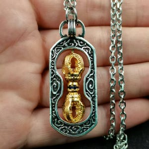 May include: A silver-toned pendant necklace featuring a gold-colored, ornate double-sided design with blue accents. The pendant is suspended from a silver chain. The pendant is rectangular with an intricate border.