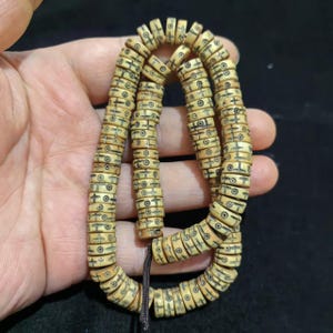 May include: A string of beige beads with black etched patterns. The beads are cylindrical with lines and circles. The string is dark brown. The beads are arranged in a spiral. Each bead is approximately 0.5 cm in diameter.
