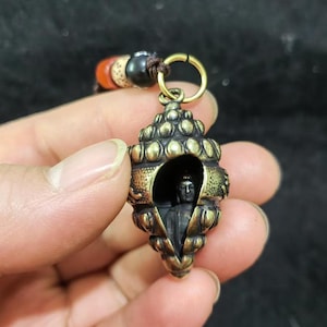 Puede incluir: Colgante de latón detallado en forma de concha, con una pequeña estatua de Buda en su interior. El colgante está unido a un anillo dorado y a una pulsera de cuentas. La concha presenta intrincados detalles y un aspecto envejecido.