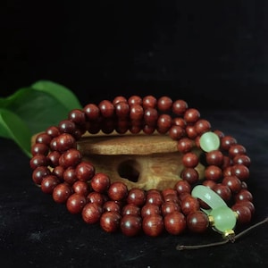 May include: A strand of reddish-brown wooden prayer beads with a light green jade accent. The beads are round and smooth, with a polished finish. The jade piece is carved into a curved shape and is attached to a brown cord.