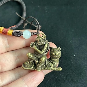 Peut inclure: Un pendentif en métal couleur laiton représentant une silhouette assise tenant un petit objet circulaire, placée à côté d'un tigre. Le pendentif est fixé à un cordon marron foncé avec une perle blanche et un accent tissé jaune et rouge.