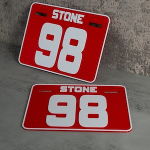 May include: Two red plastic number plates with white numbers and the word "STONE" printed on them. The top plate has the number 98 and the bottom plate has the number 98.