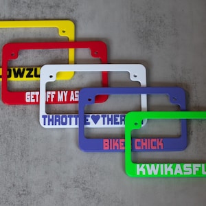 May include: Five colorful license plate frames with different phrases. The top frame is yellow with the text "SLOWZU". The second frame is red with the text "GET OFF MY AS". The third frame is white with the text "THROTTLE ♥ THER". The fourth frame is purple with the text "BIKE CHICK". The bottom frame is green with the text "KWIKASFUKI".