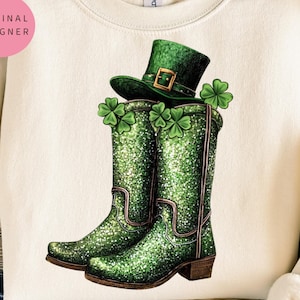以下が含まれることがあります： クリーム色のスウェットシャツに、緑色のグリッターのついたカウボーイブーツのデザインが施されています。ブーツには緑色のクローバーと緑色のレプリコーンの帽子が乗っています。