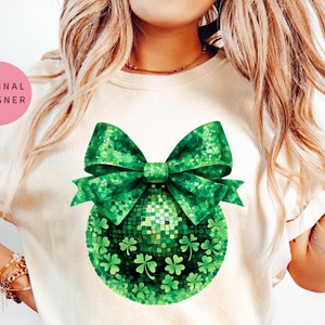Könnte beinhalten: Ein weißes T-Shirt mit einem grünen Glitzer-Kleeblatt-Anhänger und einer Schleife. Der Anhänger ist aus grünem Glitzer gefertigt und hat eine glitzernde Oberfläche.