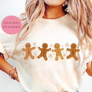 May include: A white t-shirt with a line of four gingerbread men in gold and brown with pink accents. The gingerbread men are smiling and have buttons and arms. There are snowflakes in between the gingerbread men.