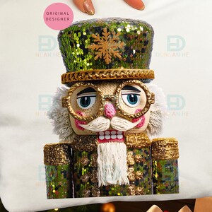 May include: A white cushion with a detailed illustration of a nutcracker. The nutcracker has a green sequined hat with a gold snowflake, gold-rimmed glasses, and a white mustache. The text "ORIGINAL DESIGNER" is in a pink circle.