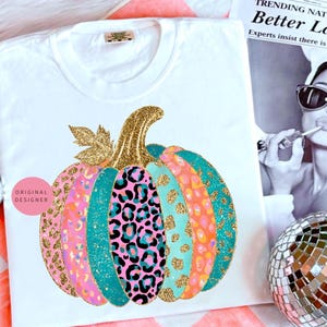 May include: White t-shirt featuring a colorful pumpkin design. The pumpkin has sections of pink, teal, orange, and leopard print patterns, with gold glitter accents. The stem and leaves are also gold glitter. The shirt has the words "ORIGINAL DESIGNER" on it.