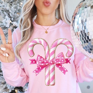 May include: Pink sweatshirt with a graphic of two candy canes tied together with a pink and white polka dot bow. The candy canes are white and pink with gold glitter accents.