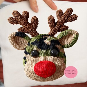 May include: A white pillow featuring a knitted reindeer design. The reindeer has a camouflage pattern head, brown antlers, black eyes, and a red nose. The ears are tan and green. The pillow is held by a hand.