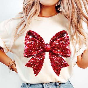 Puede incluir: Una camiseta blanca con un gran diseño de lazo rojo de lentejuelas.