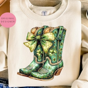 Puede incluir: Una sudadera de color crema con un diseño de dos botas de vaquero en verde y dorado con purpurina y un lazo verde. Las botas están decoradas con tréboles.