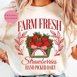 Puede incluir: Una camiseta blanca con el texto "FARM FRESH Strawberries HAND PICKED DAILY" en rojo. Una cesta de fresas rojas con un patrón de cuadros verdes y blancos y un lazo rosa se encuentra en el centro de la camiseta.