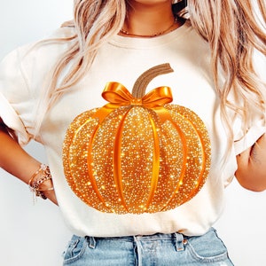 May include: A white t-shirt with a sparkly gold pumpkin graphic. The pumpkin has a brown stem and a gold bow.