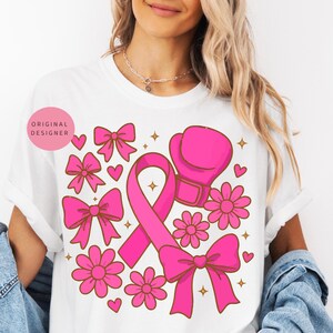 May include: White t-shirt featuring a pink ribbon, boxing glove, bows, flowers, and hearts. The design includes the text "ORIGINAL DESIGNER" in a pink circle. The graphic is in a retro style with gold star accents.