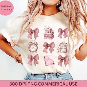 May include: A white t-shirt with a pink and white design featuring a stack of books, a clock, a backpack, a notebook, and bows. The design is perfect for teachers, students, or anyone who loves to read.