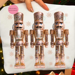 May include: Three glittery, rose gold nutcrackers with black mustaches and blue eyes are displayed on a white pillow. The nutcrackers are wearing gold and rose gold uniforms. The pillow has snowflake designs and the text "ORIGINAL DESIGNER".