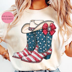 Op de afbeelding: Crèmekleurig T-shirt met een patriottisch ontwerp. De afbeelding bevat een cowboyhoed, een rode strik met een juweel en cowboylaarzen met een rood, wit en blauw Amerikaans vlagontwerp. De laarzen hebben een glittereffect.