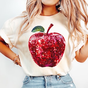 May include: A white t-shirt with a red sequined apple graphic. The apple has a green leaf and stem.