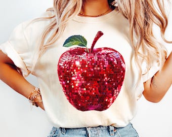 May include: A white t-shirt with a red sequined apple graphic. The apple has a green leaf and stem.