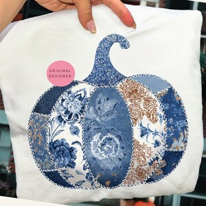 May include: White pillow with a patchwork pumpkin design. The pumpkin features blue and brown floral patterns, with a blue stem. The design is stitched onto the pillow. A pink sticker with the words "ORIGINAL DESIGNER" is visible.