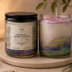 May include: Two glass jar candles with black lids sit on a round, light-colored stone. The candle on the left has a label that reads "Cereal Spectrum" and "Sense To Relax." The candle on the right has a colorful, layered wax design. The candles are surrounded by neutral decor.