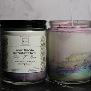 May include: Two glass jar candles. One has a black lid and a label that reads "Cereal Spectrum" and "Science To Relax". The other candle has a white wax with colorful accents of purple, green, and blue. Both candles are on a black surface.