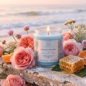 May include: A lit blue candle in a glass jar, labeled "Ocean Rose," surrounded by pink roses, orange slices, and honeycomb. The candle sits on a weathered wooden surface with a blurred beach and ocean background.