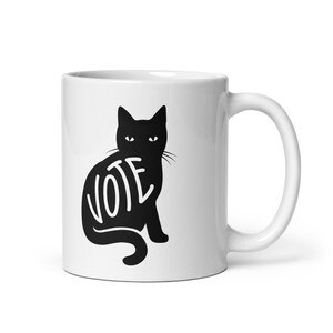 May include: White ceramic coffee mug with a black silhouette of a cat sitting with the word "VOTE" spelled out on its back.
