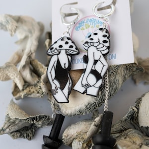 May include: A pair of silver hoop earrings with black and white mushroom-shaped charms. The charms feature a woman's silhouette in a swimsuit. The earrings have a black, stacked, cylindrical dangle. The earrings are displayed on a white card with the text "@whookearrings.com".