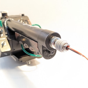 May include: A black and green handheld device with a silver tip and red and black wires extending from the tip. The device has a textured black barrel and a green wire wrapped around the barrel.