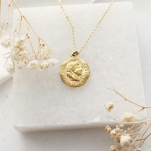 Pode incluir: Um colar com pingente de moeda dourada. O pingente apresenta um perfil e o texto "CONFOEDERATIO HELVETICA 1973". O colar &eacute; exibido em uma superf&iacute;cie de m&aacute;rmore branco, com flores secas.