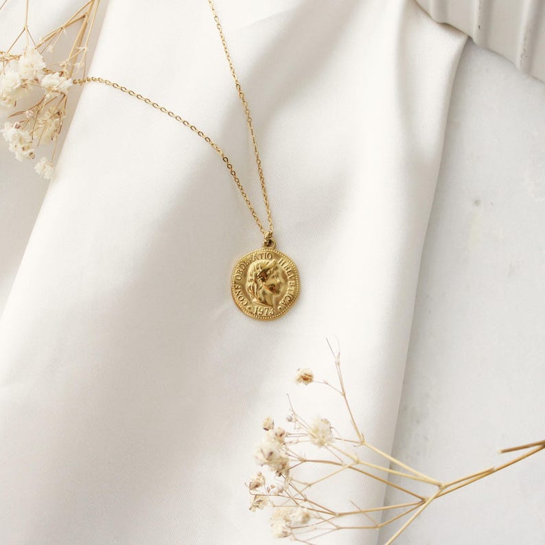 Pode incluir: Um colar com pingente de moeda dourada. O pingente apresenta um design em relevo detalhado e o ano de 1974. O colar &eacute; exibido em um fundo de tecido branco, com delicadas flores secas em primeiro plano.