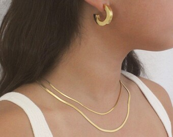 Op de afbeelding: Een gouden ketting met twee strengen, op elkaar gelegd. De ketting wordt gedragen door een persoon met lang bruin haar. De persoon draagt ook gouden oorringen.