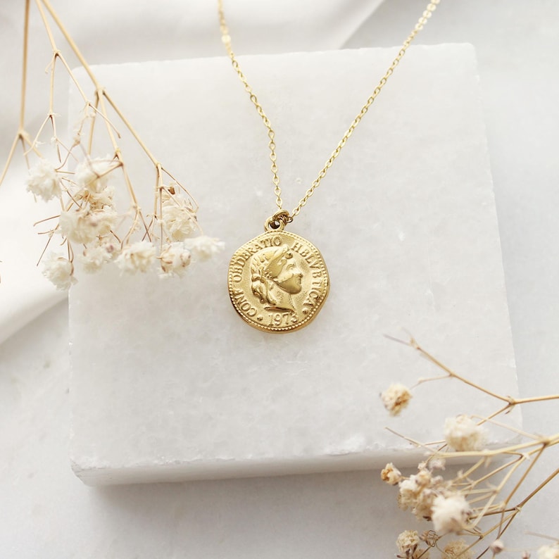 Pode incluir: Um colar com pingente de moeda dourada. O pingente apresenta um perfil e o texto "CONFOEDERATIO HELVETICA 1973". O colar &eacute; exibido em uma superf&iacute;cie de m&aacute;rmore branco, com flores secas.